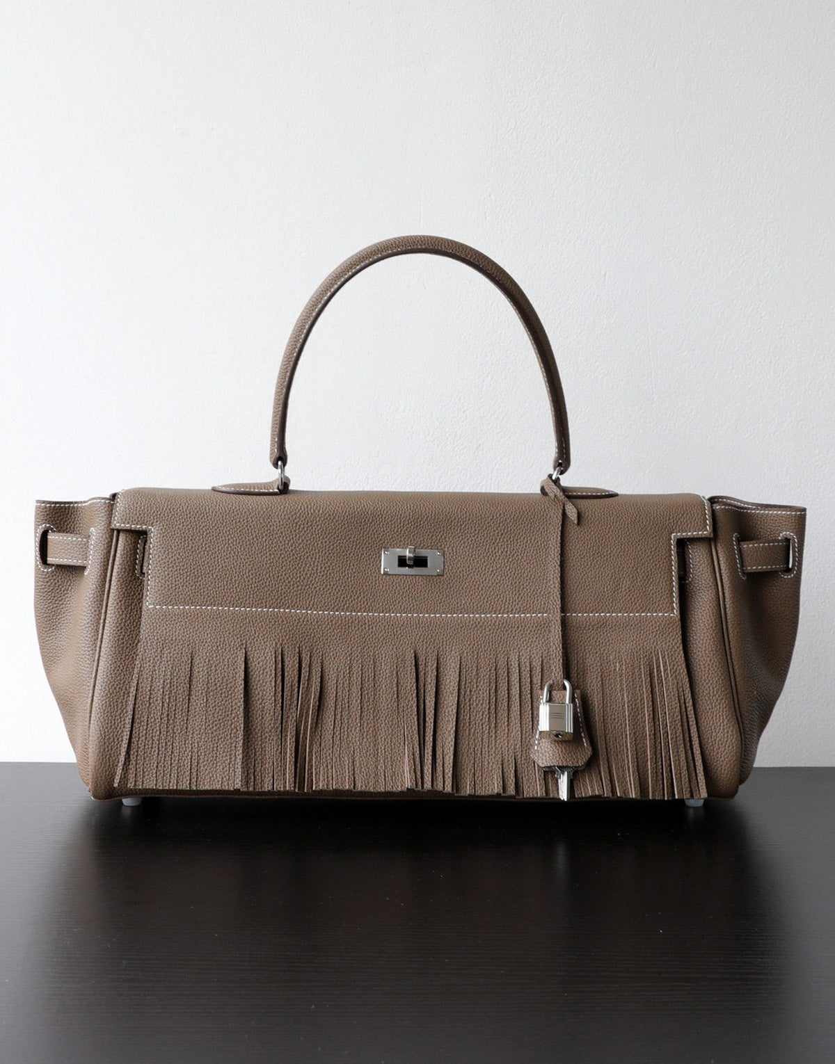 A taupe leather handbag with a fringed front detail, dual top handles, and silver-tone hardware, displayed on a dark surface against a light gray background.