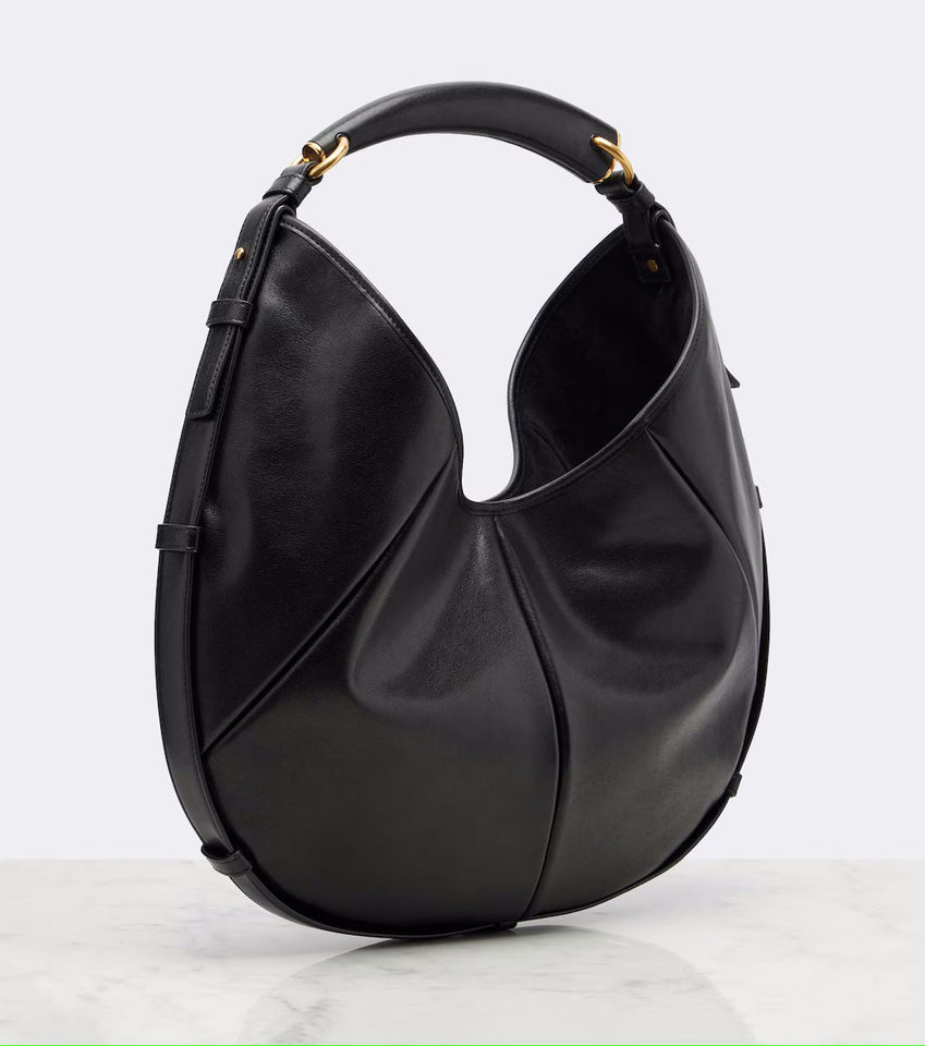 A black leather hobo shoulder bag with gold-tone hardware, displayed on a light gray background with a white marble surface.