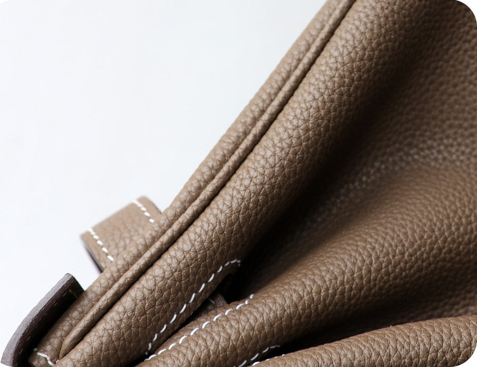 Close-up of a taupe brown pebbled leather bag corner showing white contrast stitching and a dark brown leather strap against a white background.