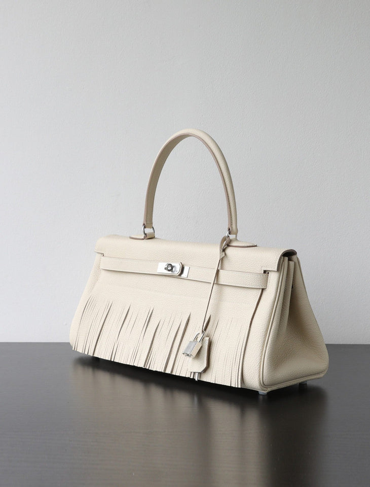 A structured cream-colored leather handbag with a distinctive fringe detail along the bottom front, a top handle, and silver-tone hardware, resting on a dark reflective surface against a light gray background.
