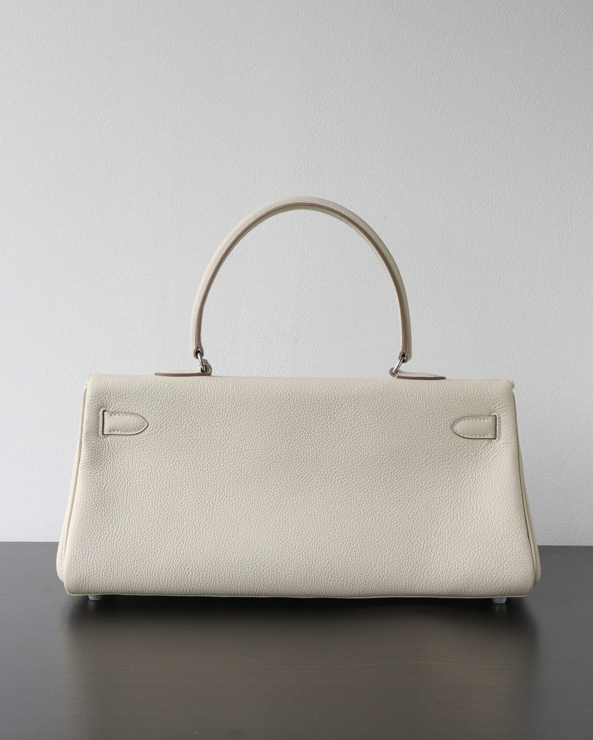 A structured cream-colored pebbled leather handbag with a top handle, resting on a dark surface against a plain light gray wall.