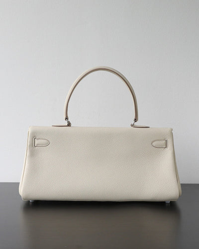 A structured cream-colored pebbled leather handbag with a top handle, resting on a dark surface against a plain light gray wall.
