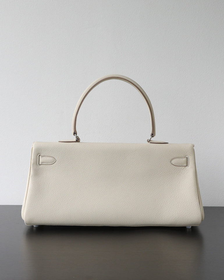 A structured cream-colored pebbled leather handbag with a top handle, resting on a dark surface against a plain light gray wall.