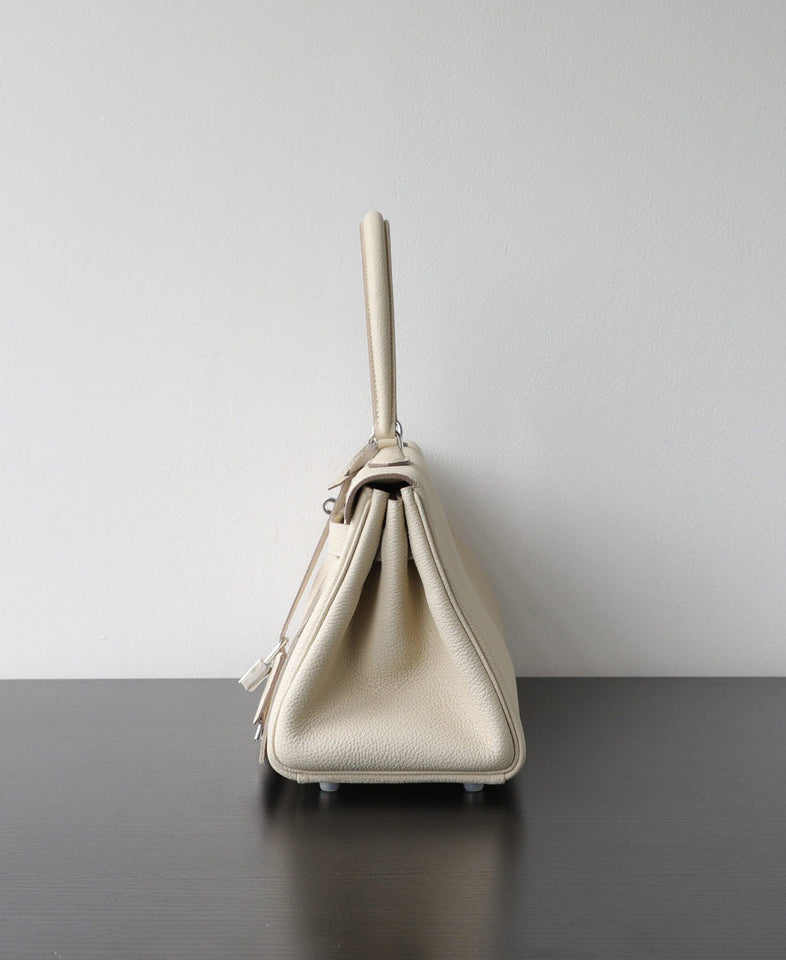 A light beige pebbled leather handbag shown from the side profile, standing on a dark wooden surface against a plain light gray wall.