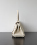 A light beige pebbled leather handbag shown from the side profile, standing on a dark wooden surface against a plain light gray wall.