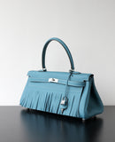 A structured blue leather handbag with a long fringe detail across the front, a top handle, and silver-tone hardware, displayed on a dark reflective surface against a light gray background.