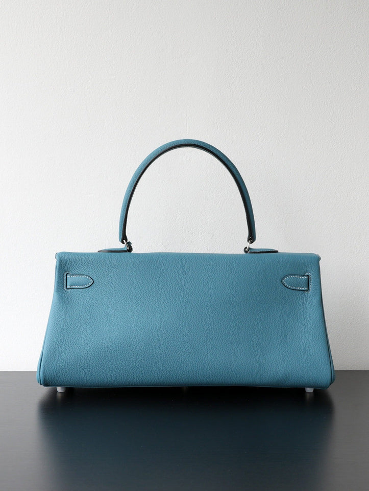 A structured top-handle handbag in textured blue leather, shown from the front on a dark surface against a plain light gray wall.