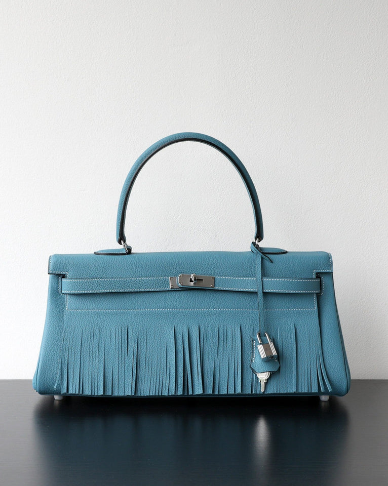 A structured turquoise blue leather handbag with a top handle, silver-tone hardware, and a long fringe detail across the front, displayed on a dark reflective surface against a plain light wall.