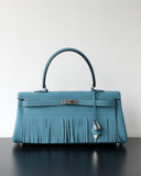 A structured turquoise blue leather handbag with a top handle, silver-tone hardware, and a long fringe detail across the front, displayed on a dark reflective surface against a plain light wall.