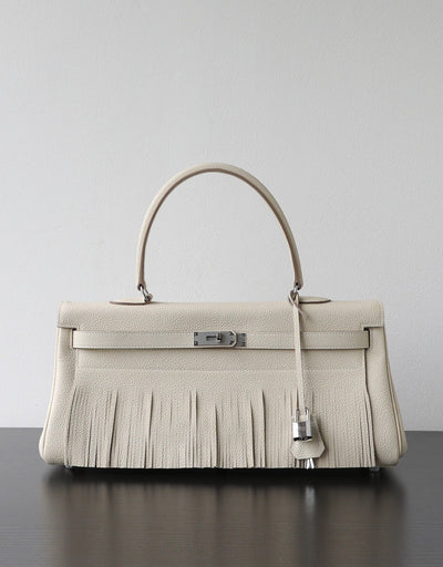 A structured cream-colored leather handbag with a fringed front, a single top handle, and silver-tone hardware, displayed on a dark surface against a light gray background.