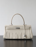 A structured cream-colored leather handbag with a fringed front, a single top handle, and silver-tone hardware, displayed on a dark surface against a light gray background.