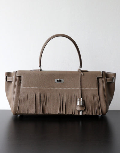 A taupe leather handbag with a fringed front detail, dual top handles, and silver-tone hardware, displayed on a dark surface against a light gray background.