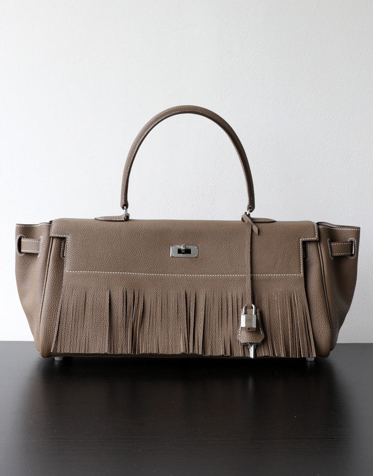 A taupe leather handbag with a fringed front detail, dual top handles, and silver-tone hardware, displayed on a dark surface against a light gray background.