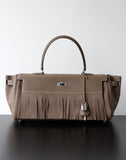 A taupe leather handbag with a fringed front detail, dual top handles, and silver-tone hardware, displayed on a dark surface against a light gray background.