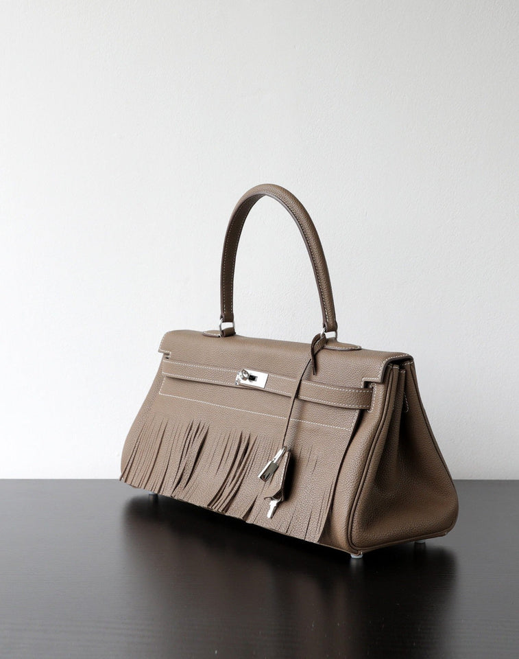 A taupe leather handbag with fringe detailing and silver-tone hardware, displayed on a dark reflective surface against a light gray background.