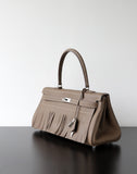 A taupe leather handbag with fringe detailing and silver-tone hardware, displayed on a dark reflective surface against a light gray background.