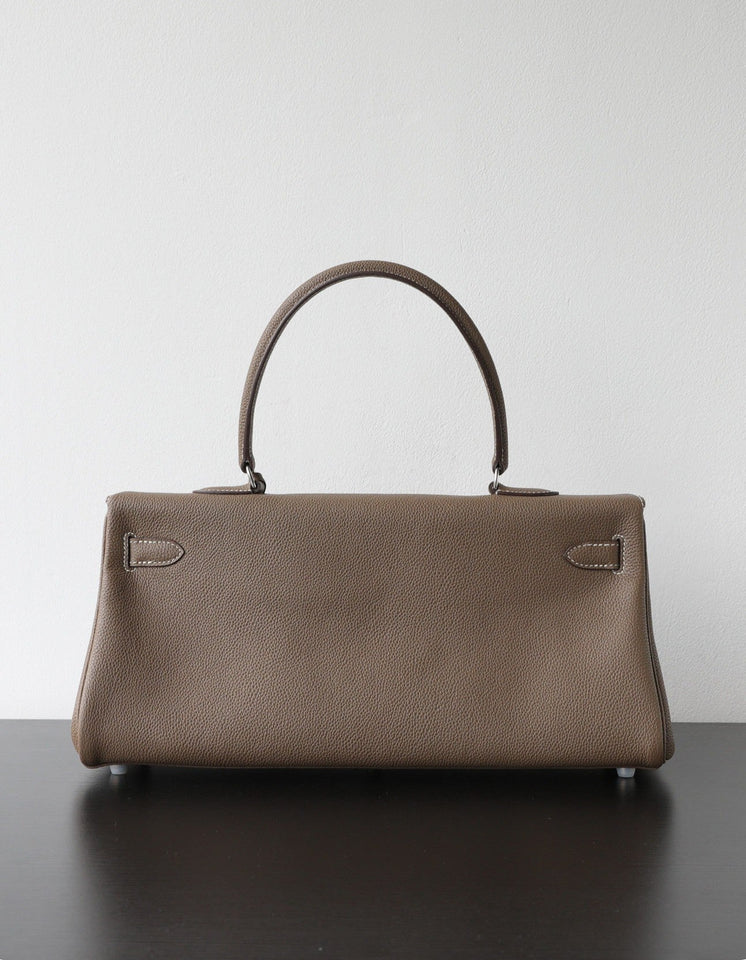 A structured taupe leather handbag with a single top handle, resting on a dark surface against a plain light gray wall.