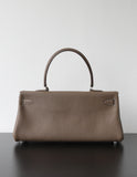 A structured taupe leather handbag with a single top handle, resting on a dark surface against a plain light gray wall.