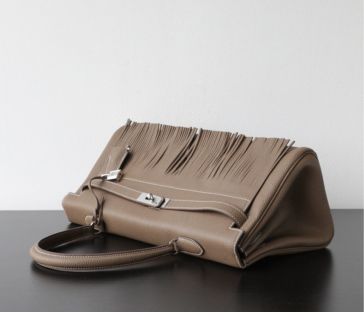 A taupe leather handbag with a distinctive fringe detail across the top flap and silver-tone hardware, resting on a dark surface against a light gray background.