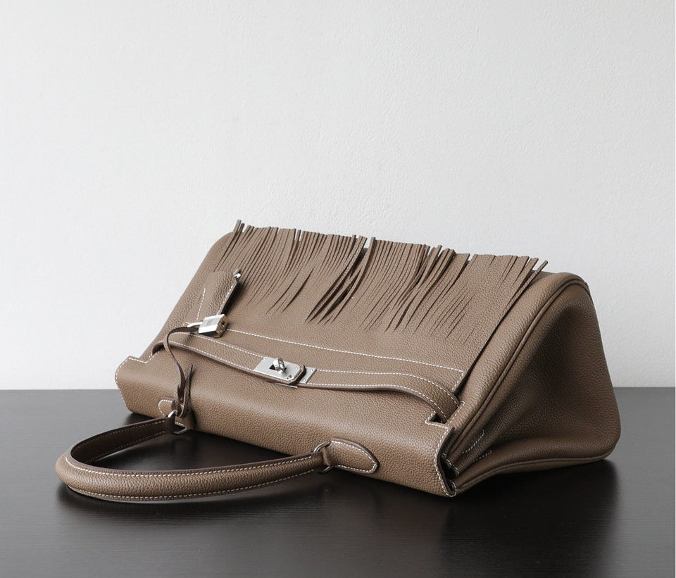 A taupe leather handbag with a distinctive fringe detail across the top flap and silver-tone hardware, resting on a dark surface against a light gray background.