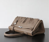 A taupe leather handbag with a distinctive fringe detail across the top flap and silver-tone hardware, resting on a dark surface against a light gray background.