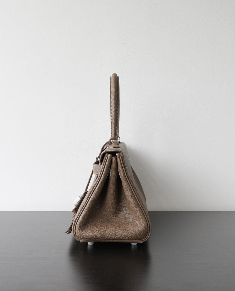 A taupe pebbled leather handbag shown from the side profile, resting on a dark surface against a plain light gray wall.