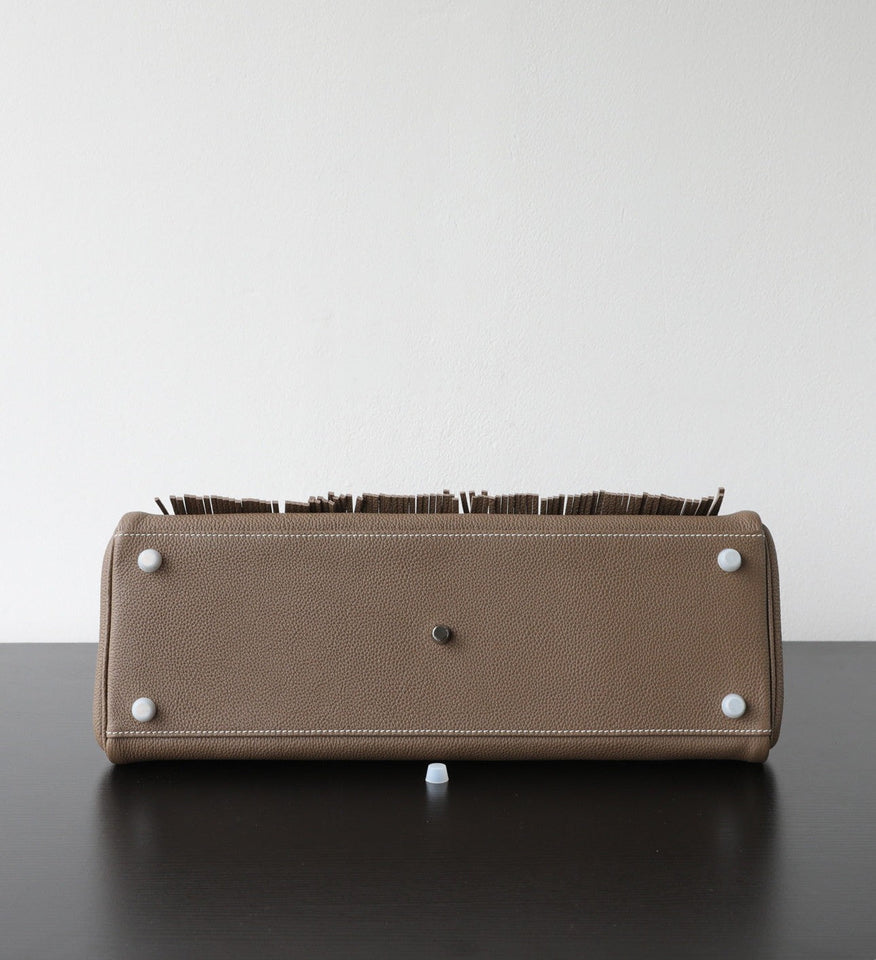 The bottom view of a taupe leather clutch or wallet with white studs and a dark brown fringe detail along the top edge, resting on a dark surface against a white background.
