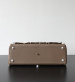The bottom view of a taupe leather clutch or wallet with white studs and a dark brown fringe detail along the top edge, resting on a dark surface against a white background.