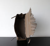 A taupe leather bucket bag with a prominent fringed flap, shown open to display its interior. The bag is positioned on a black surface against a plain white wall.
